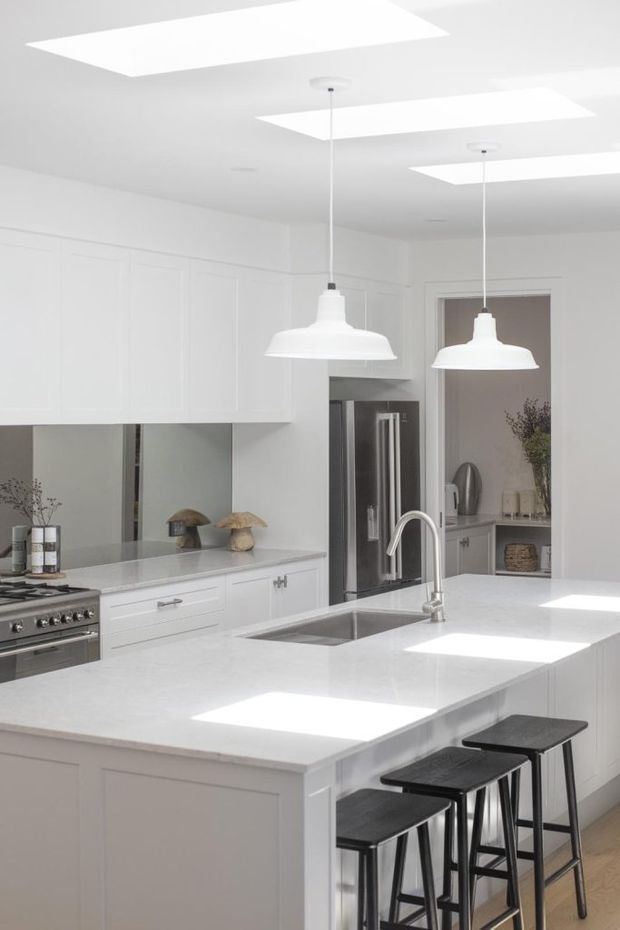 Bright, predominantly white kitchen with extensive shaker-style cabinetry, a large central island with an integrated sink and waterfall-edge benchtop, stainless-steel appliances, pendant lights, and seating for three at a breakfast bar. Skylights provide strong natural illumination, while a rear opening reveals additional kitchen storage and styling.