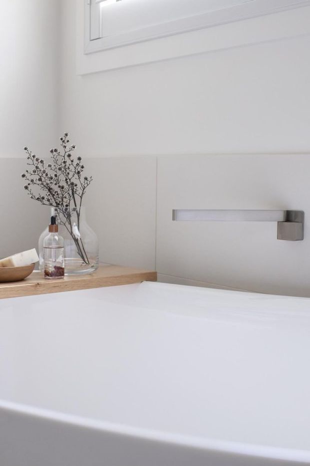 A bright, minimalist bathroom featuring a white freestanding bathtub beneath a wall-mounted rectangular chrome-silver tap. White wall panelling and trim surround a small high window, while a light timber ledge displays clear glass vessels and understated dried branches.
