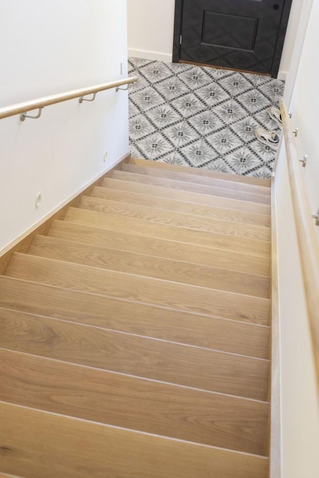A bright residential stairway viewed from above, with wide light timber-toned stair treads, white walls, simple timber handrails, and white balustrade elements. At the bottom is a patterned geometric tile landing in grey and white, leading to a dark paneled entry door.