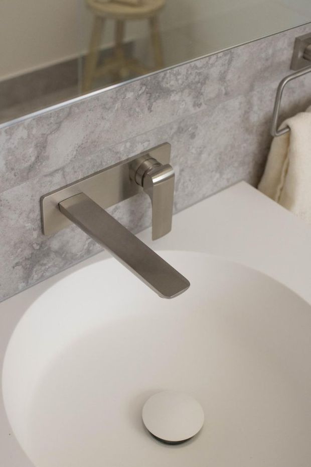 Close-up of a contemporary bathroom vanity featuring a white basin with a wall-mounted brushed metal tap and matching control. The basin is set against a pale grey, marbled stone-look tiled backsplash beneath a large mirror, with a towel rail visible at the side.