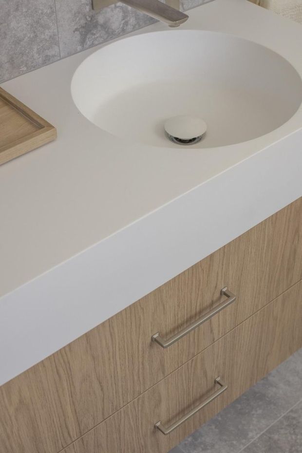 Close-up view of a contemporary bathroom vanity with an integrated oval white basin, chrome tapware and drain, pale engineered stone countertop, and light timber-look drawer cabinetry with slim metal handles. Grey stone-look wall and floor tiles frame the minimalist composition.