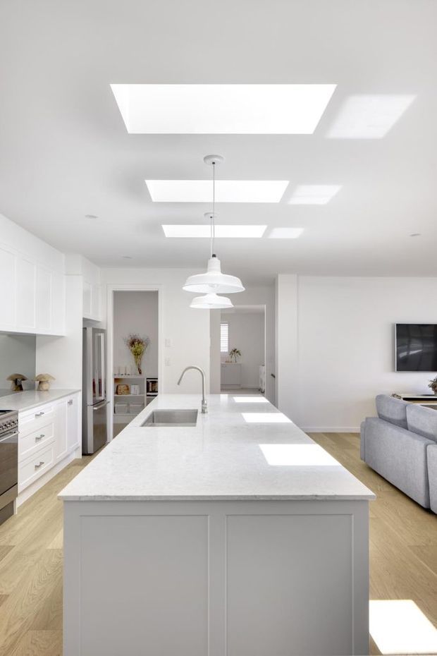 Bright open-plan white kitchen with a large central island, integrated sink and curved tap, white shaker-style cabinetry, pale stone benchtop, timber flooring, pendant lights and rectangular skylights. A living area with a grey sofa and wall-mounted television is visible beyond the kitchen.