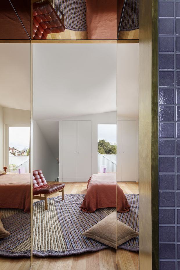 A bright contemporary bedroom viewed through full-height mirrored wardrobe panels. The room features a low bed with a rust-coloured cover, a rust leather lounge chair, a large textured striped rug, pale timber flooring, white built-in wardrobes, and a blue-purple glazed tile wall at the right. Reflections create multiple views of the room and furniture.