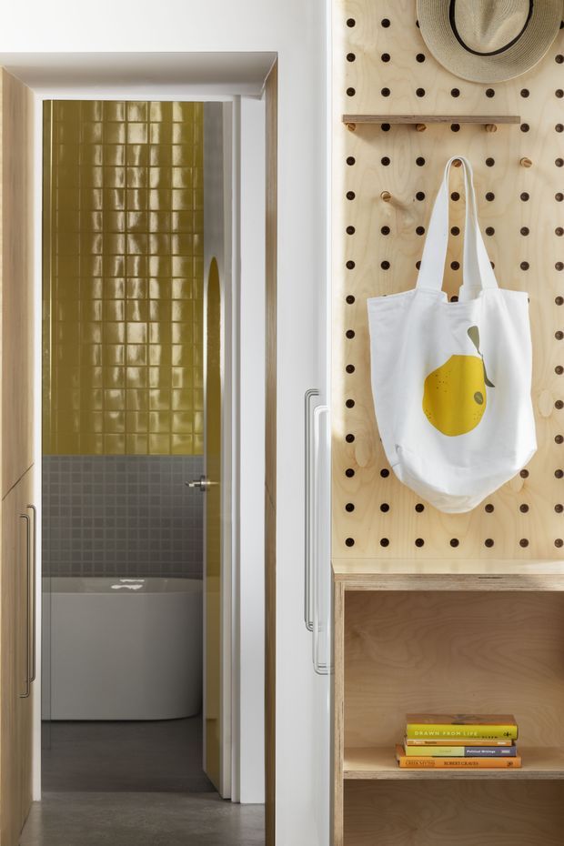 A compact, playful interior view showing a bathroom beyond a doorway and a built-in plywood pegboard storage/display wall in the foreground. The bathroom features glossy olive-green and dark grey square wall tiles, a freestanding bathtub, and dark flooring. The adjacent joinery is natural plywood with open shelving, a timber ledge, and decorative accessories including a hat and tote bag.