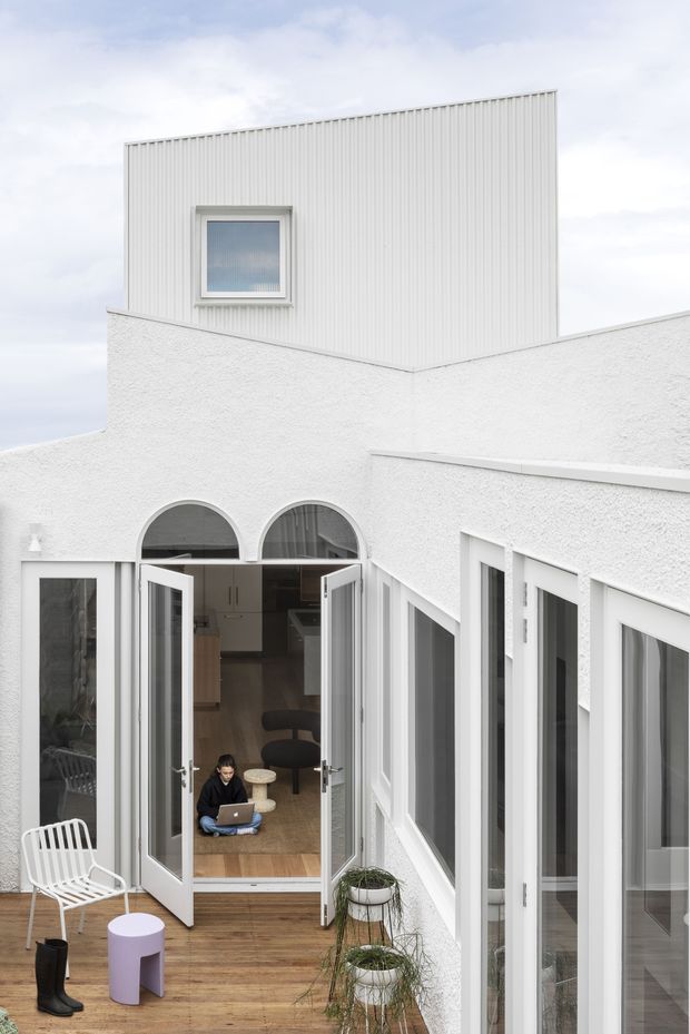 A bright white architectural courtyard viewed from above, featuring textured white rendered walls, a taller upper volume clad in vertically ribbed white panels, and extensive white-framed glazing. Folding glass doors open from the courtyard into an interior living or kitchen area. The courtyard has timber decking, potted plants, a white outdoor chair, and a small lavender side table. The composition emphasizes a crisp, minimalist contemporary renovation with strong geometric forms and abundant natural light.