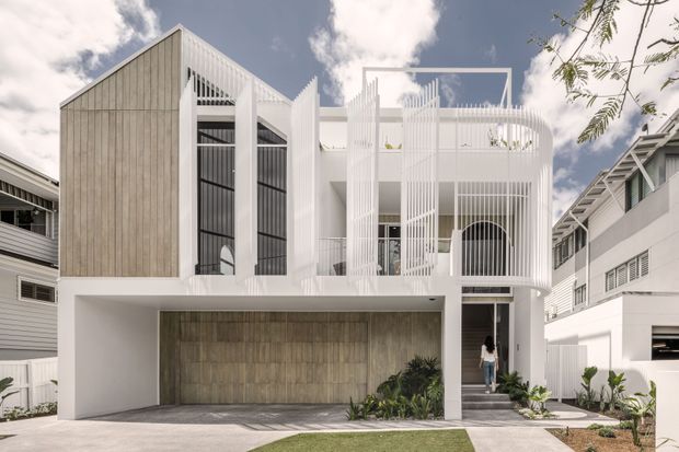 Front exterior view of a contemporary multi-level residence with a strong geometric composition. The facade combines pale vertical timber-look cladding, white rendered surfaces, tall black-framed glazing, and distinctive white vertical screening and balustrades. A covered carport or garage occupies the ground level, with a recessed entry, upper-level balcony, and landscaped front path.