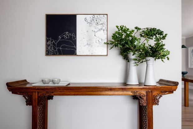 Interior view of a refined hallway or entryway featuring an ornate dark timber console table with carved detailing, decorative ceramics, white textured vases with leafy branches, and minimalist botanical artwork on a light wall. An adjoining room is partially visible at right.