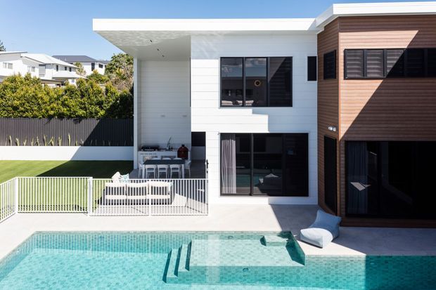Bright exterior view of a contemporary multi-level residence with a white rendered upper volume, warm timber-clad side section, extensive dark-framed glazing, and a flat roof. A landscaped courtyard includes a turquoise swimming pool, pool steps, white safety fencing, and an outdoor dining area beneath a covered terrace.