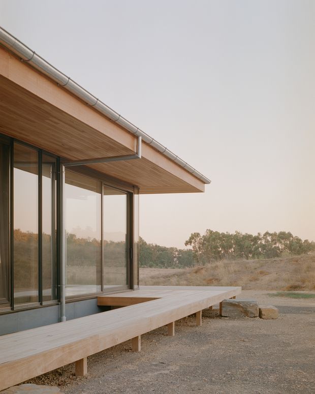 A minimalist single-storey building exterior with a broad timber-lined roof overhang, metal guttering, and full-height dark-framed glazing. A raised timber deck wraps along the facade, opening onto a dry, lightly wooded landscape. The image emphasizes the roof edge, glazing, deck, and connection between the building and its natural surroundings.