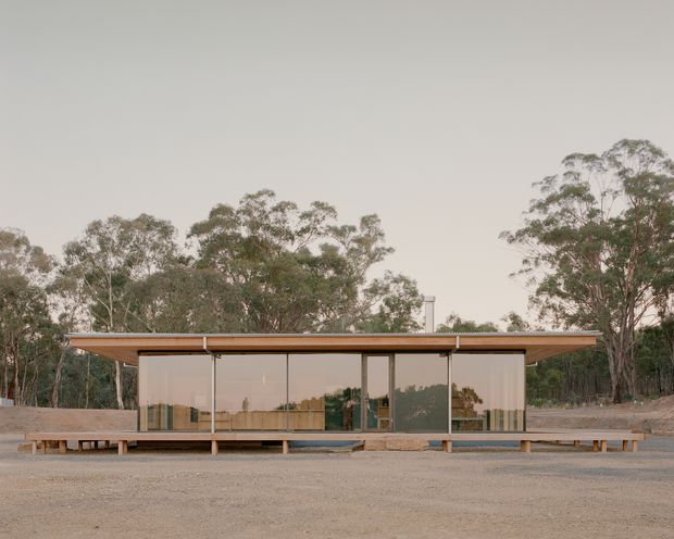 A low, single-storey modern pavilion-like building set in a eucalyptus woodland landscape. The long rectangular facade has a very thin flat roof, extensive floor-to-ceiling glazing with dark slender frames, pale vertical blinds or curtains, and a raised timber platform with simple bench-like timber elements along the front. The muted, earthy setting and restrained horizontal form create a minimalist contemporary appearance.