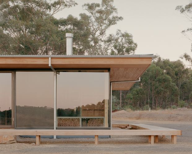 Exterior view of a low, modern pavilion-like building set among eucalyptus trees. The structure has extensive floor-to-ceiling glazing with dark metal frames, a broad shallow roof with exposed warm timber soffit, a light metal roof edge, and a simple white cylindrical chimney. A long timber bench or deck element runs across the foreground, with gravel and sparse bushland surrounding the building.