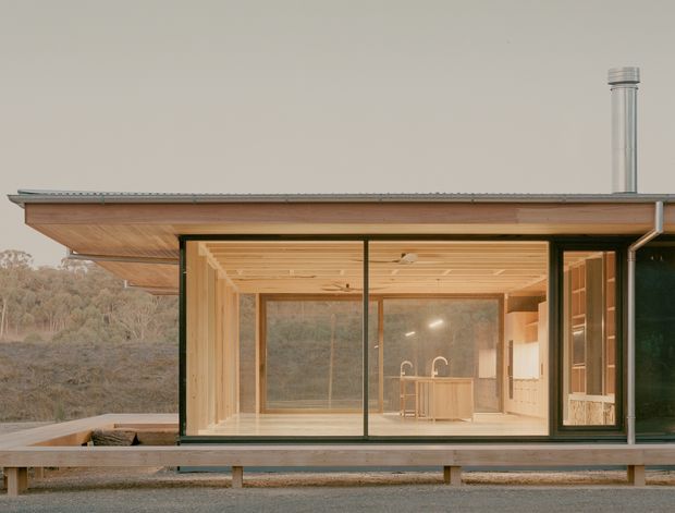 Exterior view of a compact contemporary single-storey house set in a wooded landscape. The image focuses on a broad timber-lined glazed living area with dark slender window frames, a projecting low-pitched roof, raised platform, and visible kitchen cabinetry inside. A metal flue rises through the roof, and a deck or covered outdoor platform extends along the facade.