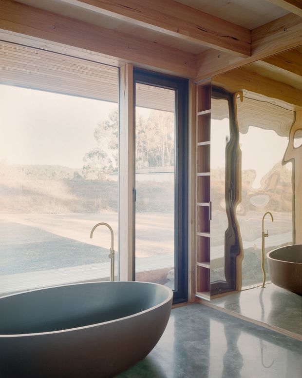 A serene, contemporary bathroom with a freestanding oval bathtub in the foreground, a sculptural wall-mounted or floor-mounted bath tap, polished grey flooring, extensive timber lining and exposed timber ceiling beams. Floor-to-ceiling glazing opens the room to a natural landscape, while a full-height reflective surface or mirror and narrow built-in shelving appear beside the window. The space has a warm, organic material palette and abundant natural light.