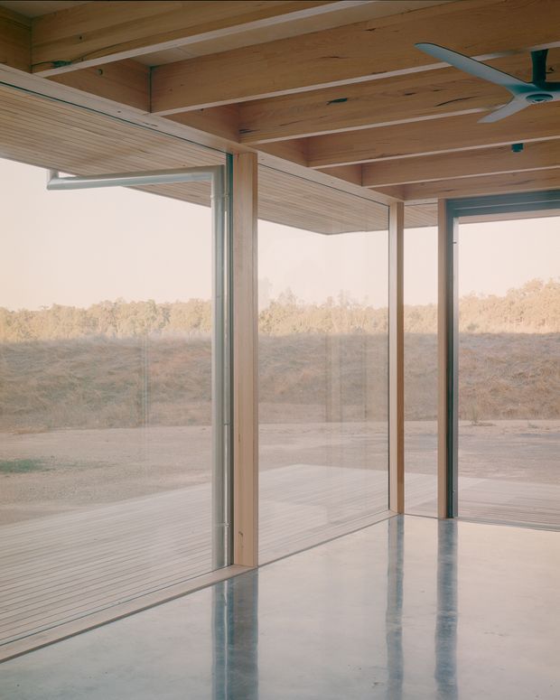 A minimalist pavilion-like interior with extensive floor-to-ceiling glazing opening onto a timber deck and dry rural landscape. Exposed light timber ceiling beams and soffit panels frame slender timber structural posts and aluminium-framed glass walls. The polished grey concrete floor reflects the glazing and ceiling, while a ceiling fan and linear light fixture provide the only visible fittings.