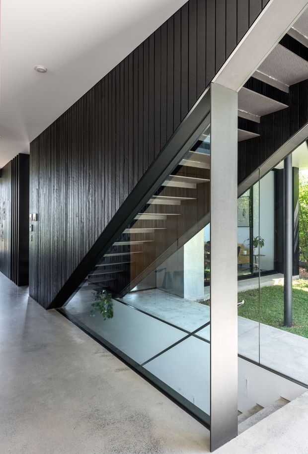 A dramatic contemporary interior stairway with open-riser timber stairs, dark vertical wall panelling, black metal framing, and extensive glass balustrades and glazing. A large pale structural column and polished concrete floor contrast with the dark stair and wall surfaces. The stairwell overlooks a lower level and connects visually to a landscaped exterior through floor-to-ceiling windows.