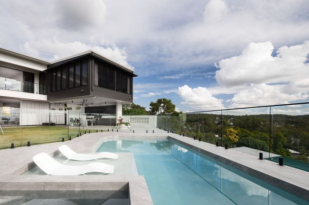 Contemporary multi-level residence with extensive glazing and a dark enclosed upper volume, overlooking a landscaped setting. The image focuses on a large elevated swimming pool with pale stone paving, integrated submerged lounge chairs, and transparent glass balustrades.
