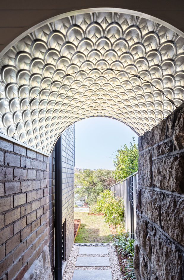 A narrow covered passageway leading from an interior or courtyard toward a garden. The focal feature is a vaulted canopy made from repeating metallic, scalloped panels that create an ornate fan-like pattern. Dark masonry walls flank the walkway, with a black metal gate and stepping stones extending toward a bright landscaped outdoor area. The image combines dramatic architectural detailing with natural daylight and greenery.