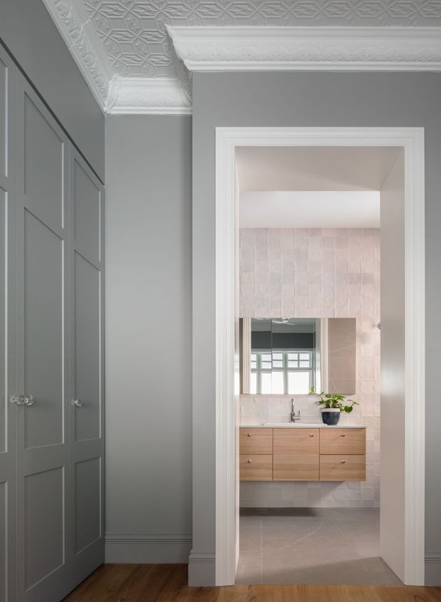 A refined transitional interior view from a hallway or dressing area into a bathroom. The foreground features tall grey-blue paneled cabinetry, dark timber flooring, white decorative pressed-metal patterned ceiling panels, and ornate white cornices and door trim. The bathroom beyond has pale blush textured wall tiles, large grey floor tiles, a floating light timber vanity with integrated drawers, a white basin, wall-mounted tapware, and a large mirror reflecting a window.