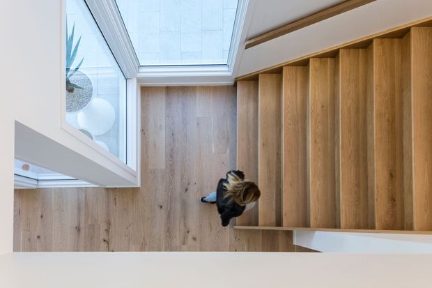 Top-down view of a refined interior stairway with warm timber flooring and matching timber stair treads, white walls, and a large skylight or glazed roof opening. A person is visible on the stairs, with a glimpse of an adjacent bright space through the glazing.