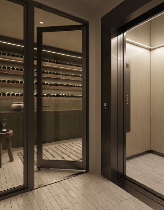 A moody, refined interior showing a compact wine room behind black-framed glass doors, with extensive horizontal wine storage and integrated warm lighting. An adjacent lift or elevator with dark framing and light wood-toned wall panels occupies the right side. The space has pale narrow-plank flooring, dark joinery, and concealed linear LED lighting.