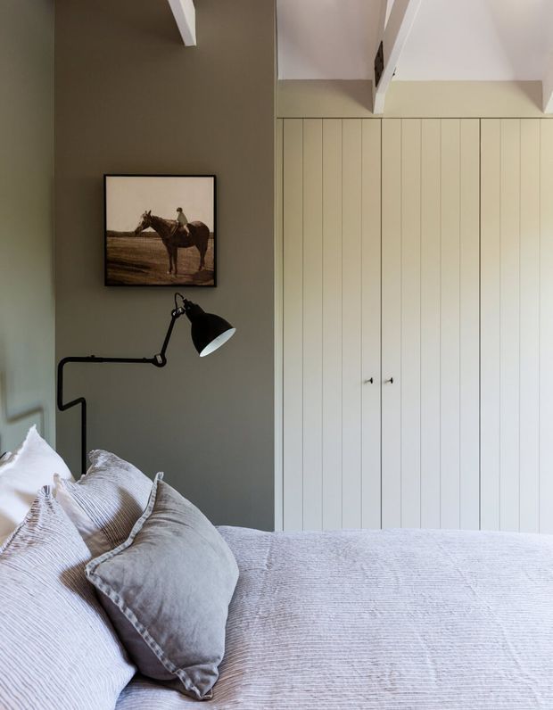 A calm, softly lit bedroom featuring a neatly made bed with textured pale bedding, a dark grey-green painted feature wall, full-height cream vertical-panel built-in wardrobes, a black articulated wall lamp, and framed equestrian artwork. White exposed ceiling beams add architectural character.