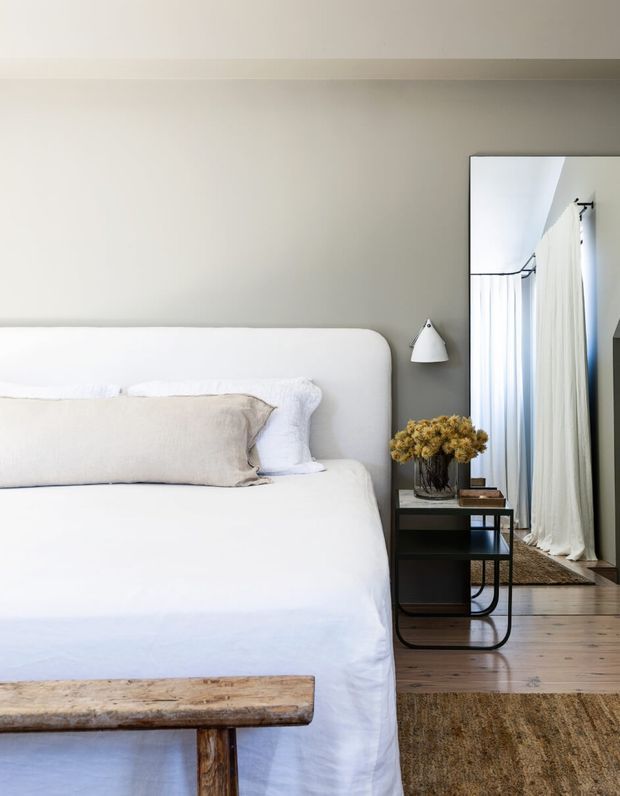 A calm, refined bedroom with a large upholstered white bed, pale linen bedding, a soft grey wall, and a slim black bedside table. A wall-mounted reading light, dried floral arrangement, timber bench, full-height mirror, sheer curtains, timber flooring, and a neutral textured rug complete the understated interior.