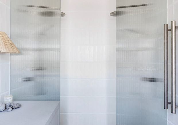 Bright, minimalist bathroom interior featuring vertically ribbed translucent glass shower panels with chrome handles, glossy white wall tiles, a pale stone vanity surface, and a partial timber-toned pendant or wall light at left.