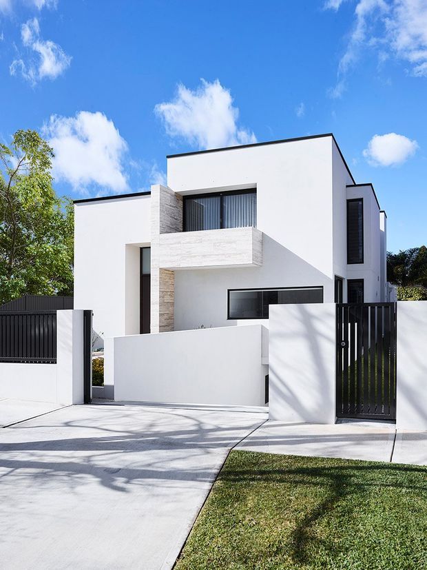 Street-facing view of a contemporary multi-level residence with crisp white rendered walls, strong rectangular volumes, dark metal-framed windows, a projecting upper balcony clad in pale stone, and a flat roofline. A white boundary wall, black vertical-slat pedestrian gate, driveway, and landscaped verge frame the facade.