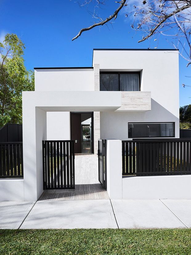 Street-facing view of a contemporary two-storey residence with crisp white rendered walls, strong cubic forms, a projecting upper-level balcony or canopy, dark aluminium-framed glazing, and black vertical metal fencing. A paved pedestrian entry leads through the front gate, with a small lawn and mature trees around the property.