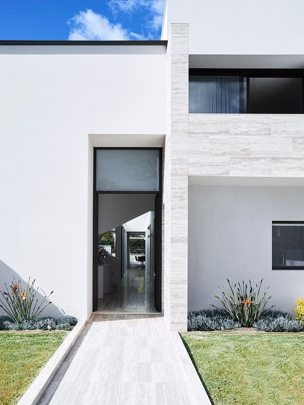 Front exterior of a contemporary two-storey residence with a crisp white rendered facade, dark-framed glazing, a tall narrow entry, and a pale stone-clad vertical feature wall. A tiled walkway leads through low, structured landscaping to the recessed entrance.