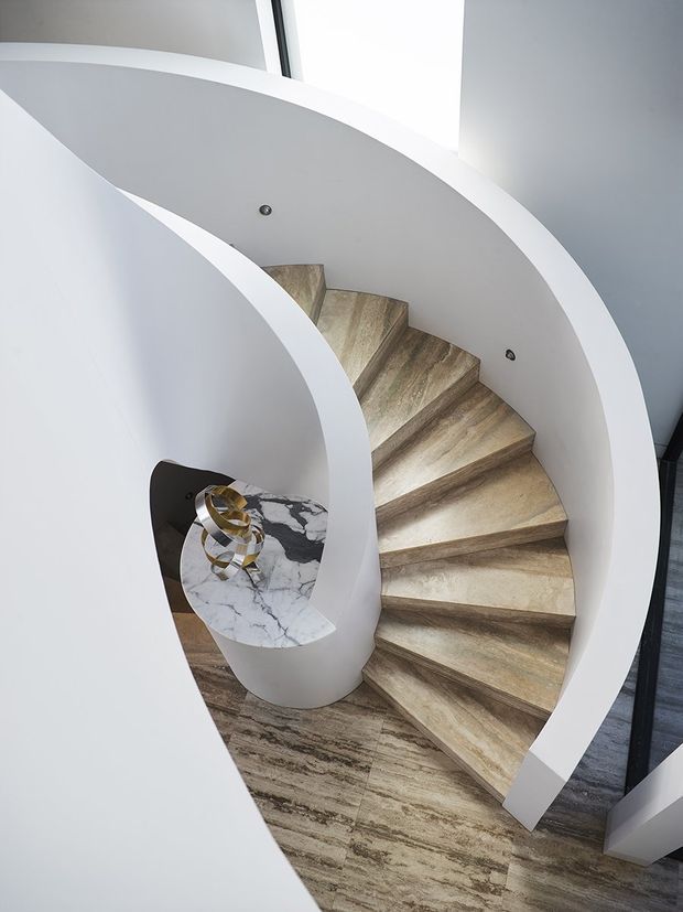 A sculptural spiral staircase viewed from above, with sweeping white curved balustrades and a central white cylindrical newel. The stair treads and surrounding floor are finished in warm, naturally patterned stone or timber-like material, while a small round marble-topped console with a gold decorative object sits beside the stair. Bright daylight enters from a high window or skylight, emphasizing the flowing contemporary form.