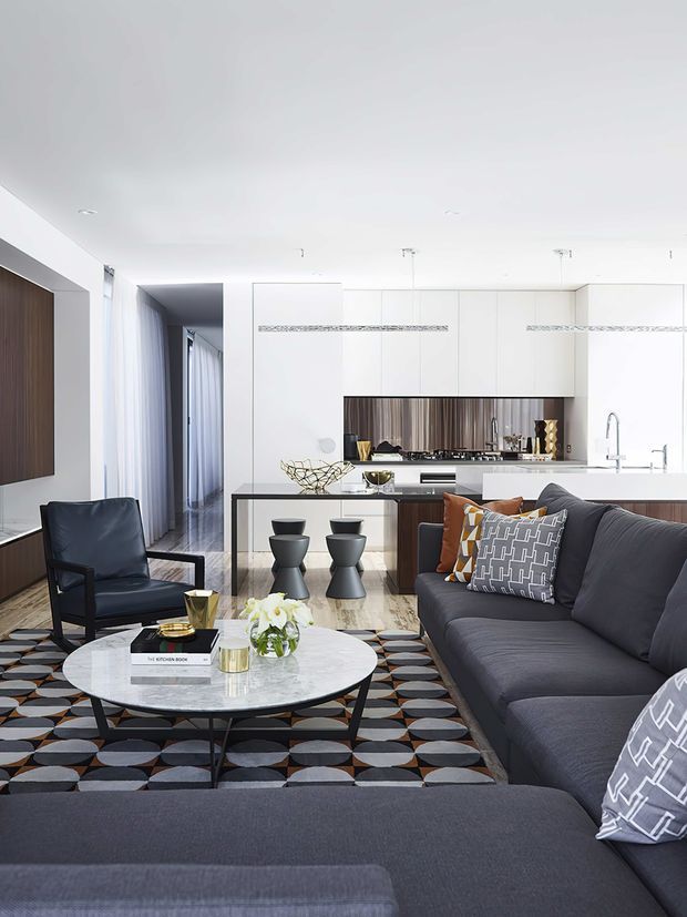 A bright open-plan living room with a contemporary kitchen in the background. The space features a large charcoal sectional sofa, black leather armchair, round marble coffee table, patterned geometric rug, sculptural side tables, and decorative cushions. The kitchen has extensive white flat-panel cabinetry, a dark timber island, a metallic textured splashback, and slim pendant lighting. A hallway and sheer curtains are visible to the left.