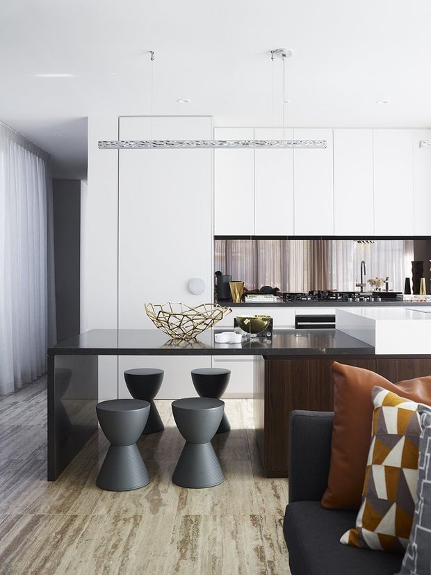 Bright contemporary open-plan kitchen and dining area with a sleek white flat-panel kitchen, dark stone island and breakfast bar, mirrored splashback, integrated appliances, and a linear pendant light. Four sculptural dark stools sit at the island, while a dark sofa with warm leather and geometric cushions occupies the foreground. Floor-to-ceiling sheer curtains and pale timber-look flooring add softness and warmth.