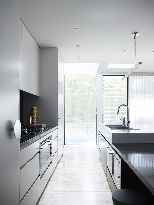 Bright contemporary kitchen with a long galley-style layout, white full-height cabinetry, dark glossy benchtops, integrated appliances, and a large central island with seating. A generous glazed door and windows bring in abundant natural light, while slim linear pendant lighting and recessed ceiling lights reinforce the clean modern aesthetic.