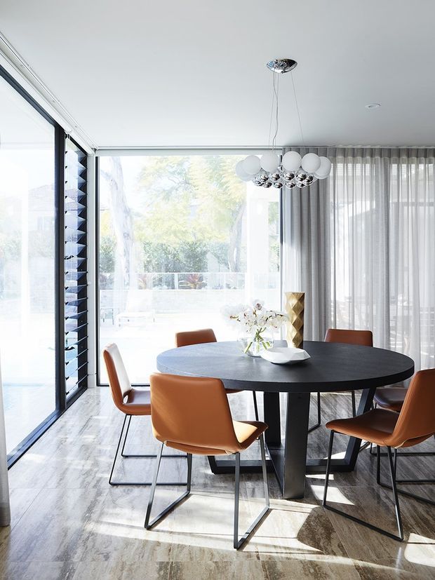 Bright contemporary dining room with a round dark dining table and tan leather chairs, surrounded by expansive floor-to-ceiling glazing and sheer curtains. The room features a sculptural spherical pendant light, pale stone-look flooring, and a light neutral palette with strong natural daylight.