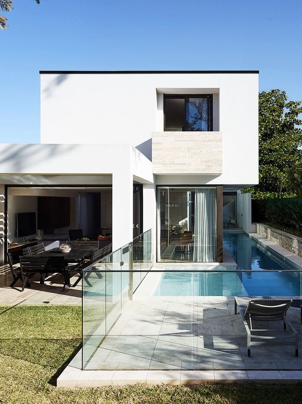 Contemporary white two-storey house viewed from the rear, with clean rectilinear volumes, large glazed openings, a projecting stone-clad upper balcony, and a private swimming pool alongside a paved terrace. A covered outdoor dining area opens directly onto the pool zone, with frameless glass balustrades and dark-framed glazing. Bright sunny setting with landscaped garden surroundings.