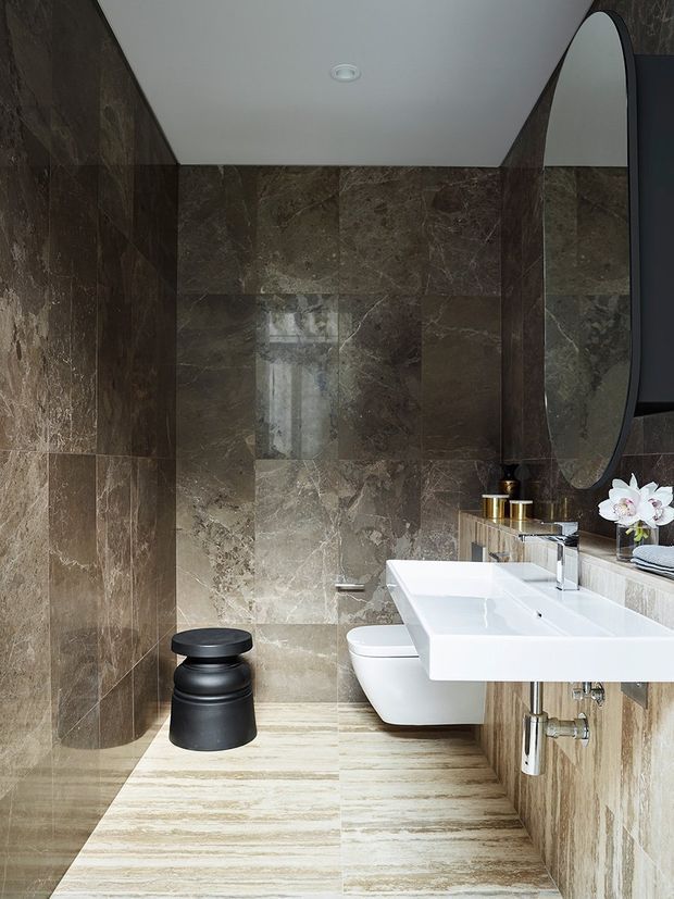 A refined contemporary bathroom featuring large-format dark brown and grey veined stone tiles on the walls, a pale striped stone floor, a floating white double vanity with integrated basins, wall-mounted chrome tapware, a wall-hung toilet, and a large oval mirror. A small black sculptural stool and metallic accessories add contrast.
