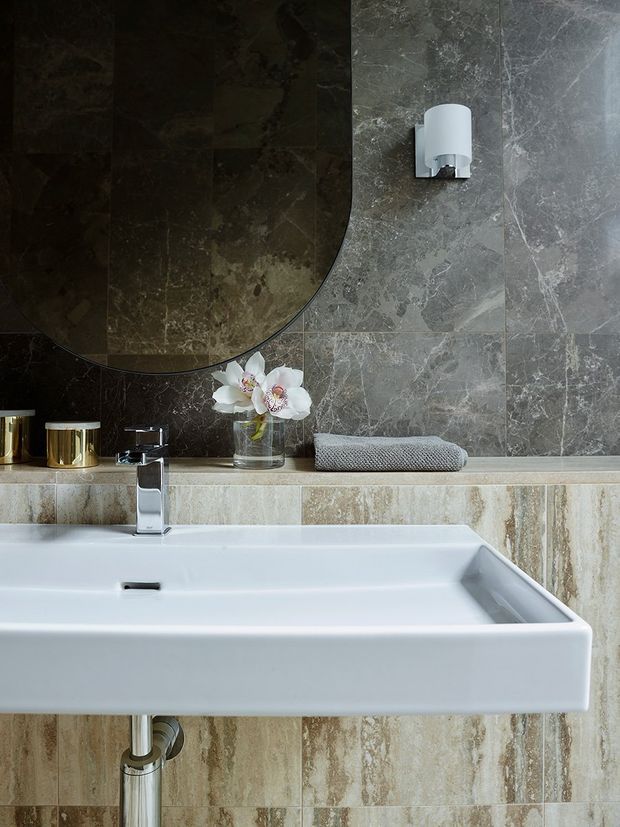 Close-up view of a refined bathroom vanity with a large white rectangular basin, chrome wall-mounted faucet, exposed chrome plumbing, and a grey stone tiled wall. A dark rounded-edge mirror, wall light, folded towel, orchid, and metallic accessories complete the scene. Beige travertine-style tiles form the vanity backsplash and lower wall, creating a luxurious, moody finish.