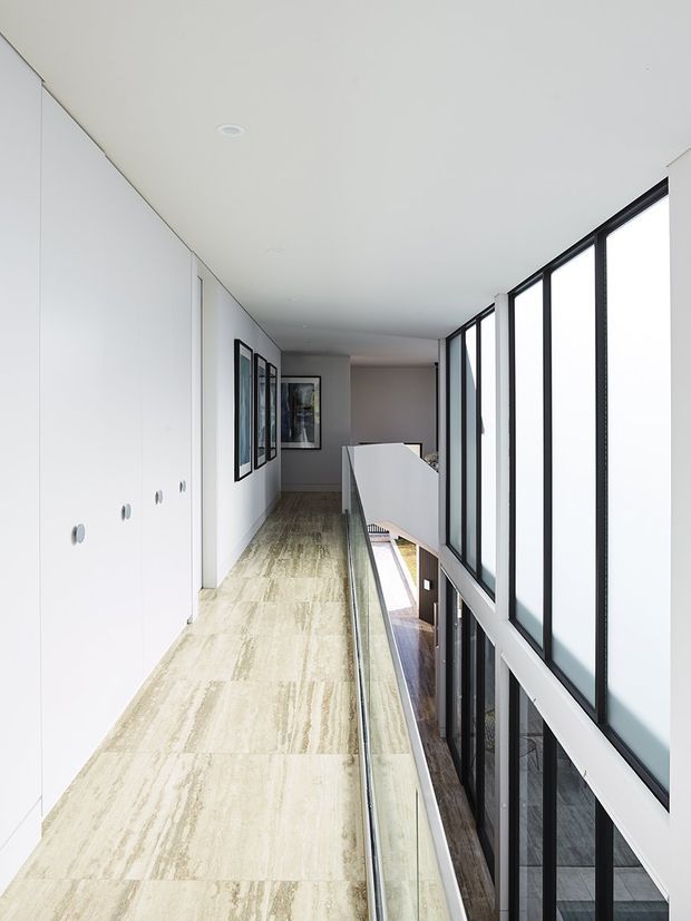 A bright, minimalist upper-level hallway or landing with large black-framed windows along one side overlooking a lower floor. The corridor has pale stone or concrete-look flooring, white built-in storage cabinetry, white walls and ceiling, framed artwork, and a clear glass balustrade. The space is clean, elongated and contemporary, with strong natural light and a double-height void visible beyond the railing.