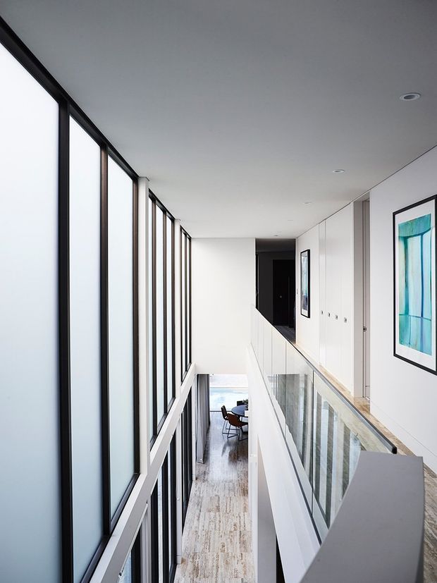 Bright contemporary interior corridor or landing overlooking a lower-level dining area. Full-height black-framed glazing lines the left side, while a glass balustrade and white built-in cabinetry run along the right. White walls and ceiling, pale timber flooring, recessed downlights, and minimalist framed artwork create a clean, airy double-height space.