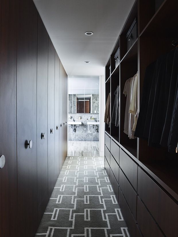 A narrow, elongated walk-in wardrobe or dressing corridor with dark timber built-in cabinetry on both sides. The left side has tall closed wardrobe doors with round knobs, while the right side has open hanging rails, drawers, and folded clothing storage. A patterned grey carpet runs along the floor toward a bright bathroom at the rear, which features a large mirror, double vanity, and grey stone finishes. Recessed ceiling lights provide even illumination, creating a refined, moody, high-contrast interior.