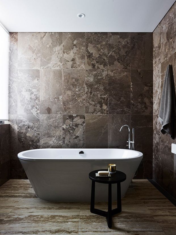 A sophisticated bathroom featuring a freestanding white bathtub against walls clad in large-format dark brown and taupe natural stone tiles. The room has matching stone-look flooring, a white ceiling with recessed lighting, a chrome floor-mounted bath filler, and a small black side table with toiletries. A dark towel hangs on the right wall.