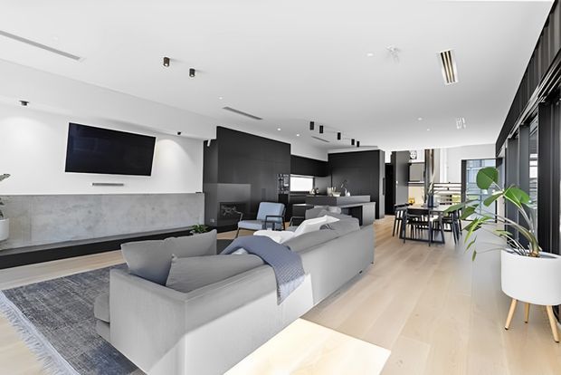 A bright, open-plan contemporary living space with a large pale sectional sofa facing a wall-mounted television and long grey stone fireplace surround. A dark kitchen with a central island is visible beyond the lounge, while a dining table sits beside full-height glazing and dark vertical window screening. Pale timber flooring, white walls, recessed lighting, and a large indoor plant create a clean, refined interior.