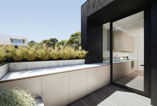 Modern residential courtyard terrace with dark timber-clad framing and large black-framed glazing opening into a bright minimalist kitchen. A long integrated pale stone or concrete bench with concealed cabinetry borders the planted garden bed, while timber decking extends across the outdoor space. The composition emphasizes indoor-outdoor connection, clean geometric lines, and restrained natural materials.