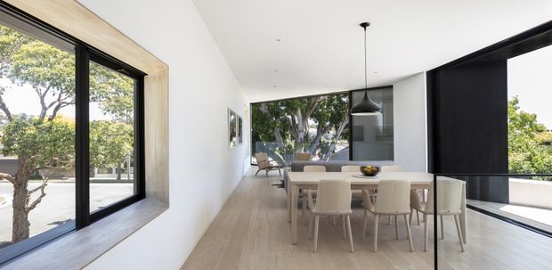 Bright contemporary open-plan dining area with a long pale timber dining table and upholstered chairs, surrounded by extensive black-framed glazing. White walls, pale timber flooring, a sculptural black pendant light, and views to mature trees create a clean, airy interior connected to an outdoor terrace.