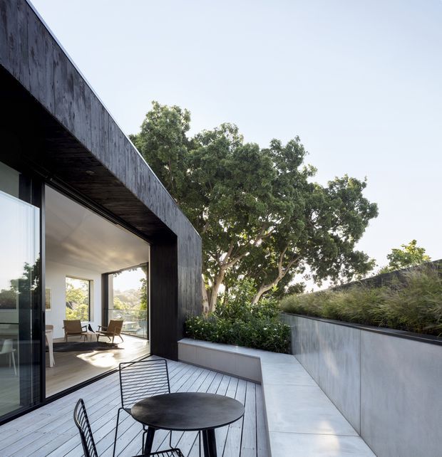 Modern architectural exterior view of a refined patio terrace beside a glazed living space. The composition features a dark timber-clad flat roof and deep overhang, full-height glazing, pale timber decking, a concrete planter and retaining wall, and built-in greenery. Outdoor dining furniture occupies the foreground, while a bright interior living area is visible through the open glazing.