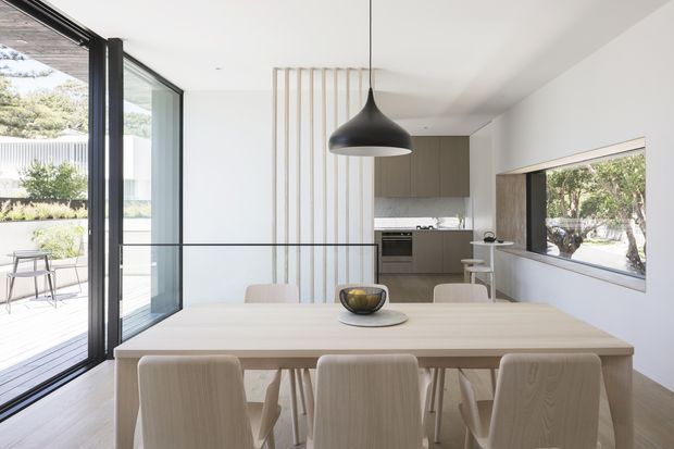 Bright contemporary open-plan dining and kitchen interior with a large pale timber dining table and matching chairs in the foreground. Full-height black-framed glazing opens toward an outdoor deck, while a large horizontal window brings in views of trees. The kitchen beyond has muted grey-beige cabinetry, a dark benchtop, integrated appliances and a vertical timber-slat feature screen. A sculptural black pendant hangs above the dining table.