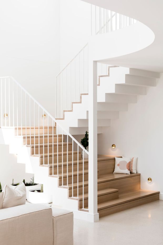 Bright, minimalist double-height interior centred on a sculptural white staircase with slim white metal balustrades and warm timber treads. A second stair flight forms built-in timber seating and drawers beneath it, with integrated shelving, cushions, small wall lights and potted plants. A pale upholstered sofa occupies the foreground, and a curved stair or landing edge creates a strong architectural feature.