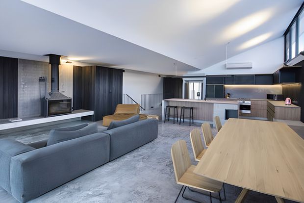 A spacious open-plan living, dining and kitchen area with a contemporary fit-out. The room features a large grey sectional sofa, timber dining table with matching chairs, a central kitchen island with bar seating, dark built-in cabinetry, polished concrete-style flooring, and high clerestory glazing. A black cylindrical fireplace anchors the living area, while a stair and entry zone are visible in the background.