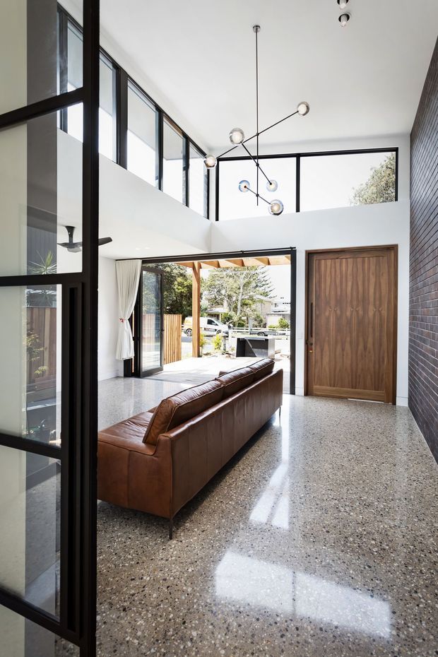 A bright, double-height residential entry and living space with polished terrazzo flooring, black-framed glazing, a large timber front door, exposed dark brickwork, and a brown leather sofa. A sculptural multi-arm pendant hangs above the open-plan area, with outdoor landscaping visible through the open doors.